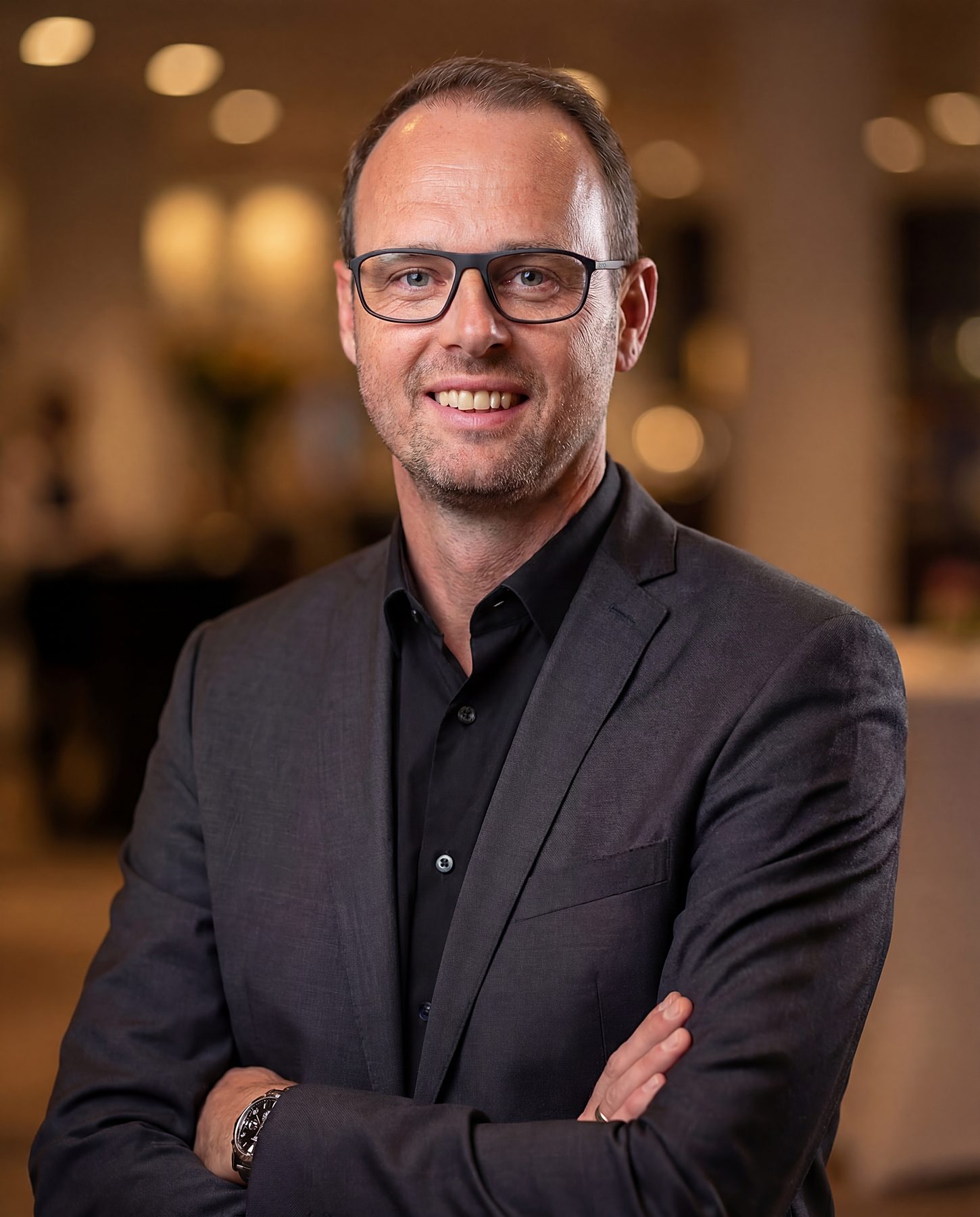 Porträt von David Beike: dunkles Hemd, dunkles Sakko, verschränkte Arme, ein offenes Lächeln